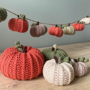 May include: A garland of crocheted pumpkins in various shades of green, orange, and brown hangs from a string.  There are also five larger crocheted pumpkins on a wooden surface, including a large orange pumpkin, a smaller green pumpkin, and two smaller cream-colored pumpkins.