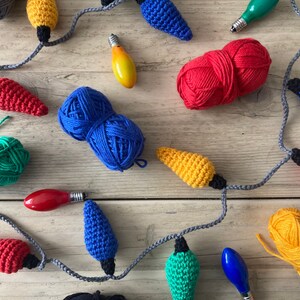 May include: A string of crocheted lightbulb ornaments in various colors, including red, yellow, blue, and green. The ornaments are strung together with gray yarn and are displayed on a wooden surface. There are also balls of yarn in various colors, including red, blue, yellow, and green.