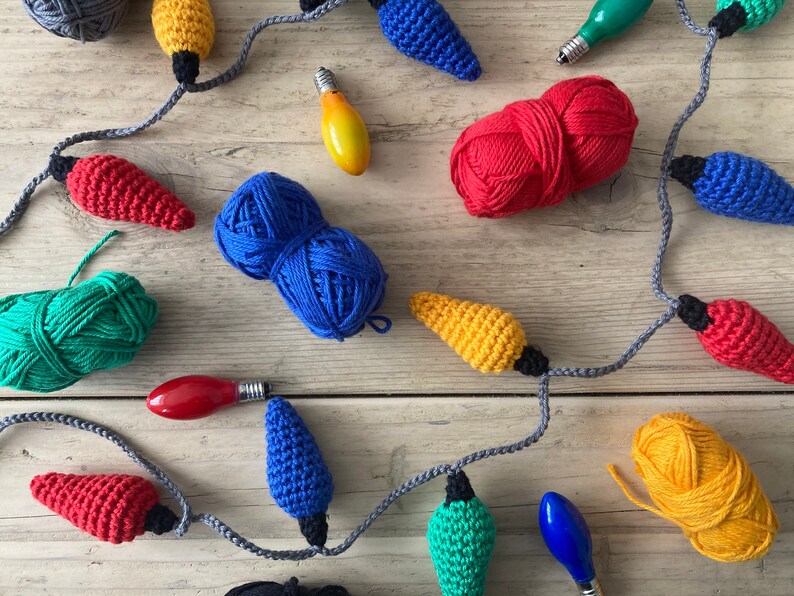 May include: A string of crocheted lightbulb ornaments in various colors, including red, yellow, blue, and green. The ornaments are strung together with gray yarn and are displayed on a wooden surface. There are also balls of yarn in various colors, including red, blue, yellow, and green.