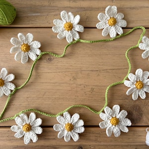 May include: A handcrafted crochet daisy garland featuring white petals and yellow centres, strung on a green vine. The garland is displayed on a wooden surface, with balls of yellow and green yarn, and a ball of white yarn in the background. The daisy garland is a decorative item.