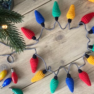 May include: A string of crocheted Christmas lights in red, yellow, blue, and green. The lights are connected by a gray cord and are arranged in a circle on a wooden surface.