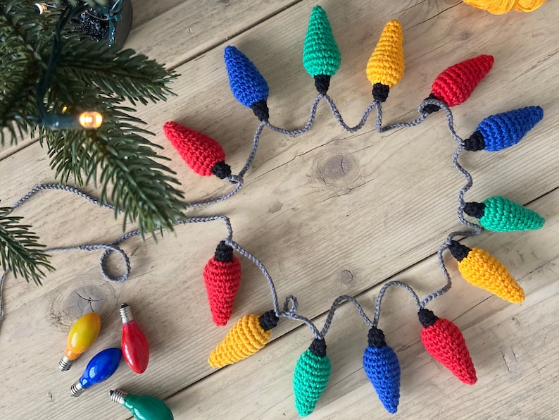 May include: A string of crocheted Christmas lights in red, yellow, blue, and green. The lights are connected by a gray cord and are arranged in a circle on a wooden surface.