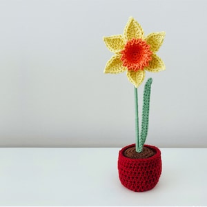 May include: A crocheted daffodil flower in a red pot. The flower is yellow with an orange centre and green leaves.