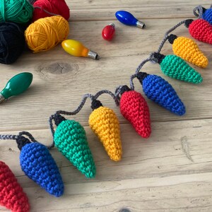 May include: A string of crocheted light bulbs in various colors, including red, yellow, green, and blue. The bulbs are connected with gray string and are arranged in a line on a wooden surface.
