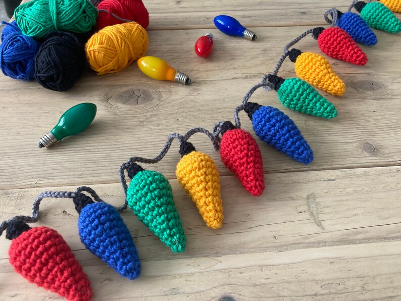 May include: A string of crocheted light bulbs in various colors, including red, yellow, green, and blue. The bulbs are connected with gray string and are arranged in a line on a wooden surface.