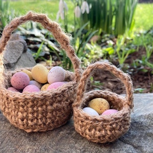 May include: Two small brown jute baskets filled with pastel colored chocolate eggs. The baskets are sitting on a rock with green grass and white flowers in the background.
