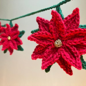 以下が含まれることがあります： ポインセチアの花と金のアクセントが付いた、赤と緑のクロシェ編みガーランド。ガーランドは、ホリデーシーズンに飾るのに最適です。