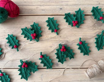 May include: A handmade Christmas garland featuring crocheted green holly leaves with red berry clusters. The garland is strung on twine and rests on a wooden surface, with green and red yarn balls in the background. A festive holiday decoration.