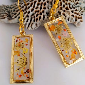 Può includere: Orecchini rettangolari dorati con fiori secchi e glitter incapsulati in resina trasparente. Gli orecchini hanno chiusura a gancio dorata. Sfondo bianco e corallo strutturato.