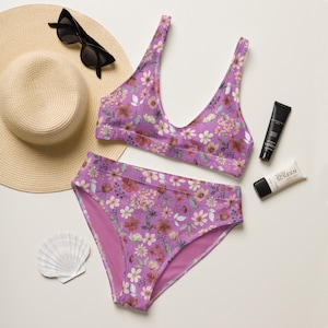 May include: A purple floral print bikini with a halter top and high-waisted bottoms. The bikini is on a white background with a straw hat, sunglasses, and sunscreen.