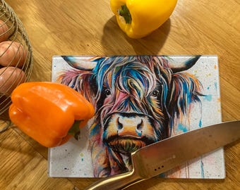 Puede incluir: Una tabla de cortar de vidrio colorida con una pintura de una vaca de las tierras altas. La vaca tiene una larga y fluida melena de pelo azul, rosa y morado. La tabla está sobre una superficie de madera con un cuchillo y dos pimientos.