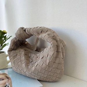 May include: A beige handbag featuring a woven texture throughout the exterior. The bag has a soft, rounded shape with a knotted handle integrated into the top design. The material appears to have a matte, suede-like finish with a consistent light tan color.
