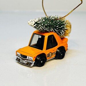 May include: Orange toy car Christmas ornament with a small green and white Christmas tree on top. The car has black wheels and a gold string for hanging. The ornament has the number 23 on the side.