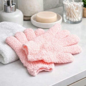 May include: A pair of light pink exfoliating gloves made of a textured material. The gloves are lying on a white surface next to a white towel, soap, and other bathroom accessories.
