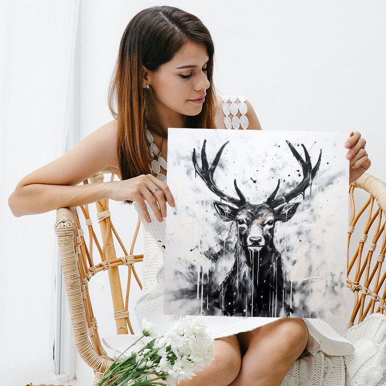 May include: A woman holds a black and white painting of a deer with large antlers. The deer is looking directly at the viewer. The painting is on a white background with black splatters of paint.