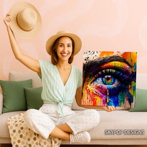 May include: A colorful square canvas print featuring a close-up of a human eye. The eye is depicted with vibrant rainbow colors, with paint drips and splatters. The print is held by a person wearing a straw hat and a light green top.