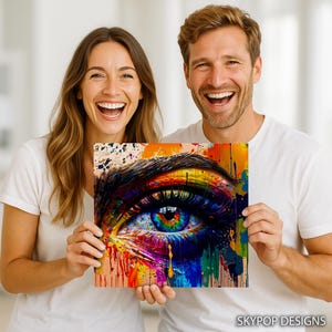 May include: A vibrant, square art print featuring a close-up of a human eye. The iris is a rainbow of colors, with blue, yellow, and purple hues. The artwork has a paint-splatter effect, with drips of color cascading down. The print is held by two people.