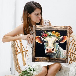 May include: A framed painting of a white cow with a colorful flower crown. The cow is looking directly at the viewer with a sweet expression. The frame is made of weathered wood with a rustic look.