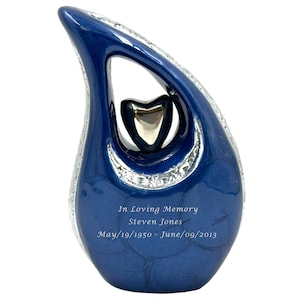 Peut inclure: Une urne en céramique bleue avec une découpe en forme de cœur argenté et des accents argentés. L'urne porte l'inscription "In Loving Memory Steven Jones May/19/1950 - June/09/2013".