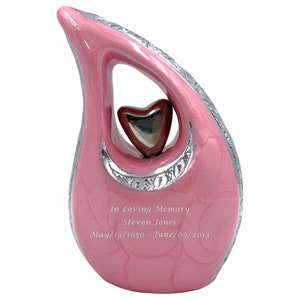 Puede incluir: Urna de cerámica rosa con un corazón recortado en plata y un borde plateado. La urna tiene la inscripción "In Loving Memory Steven Jones May/19/1950-June/09/2013".