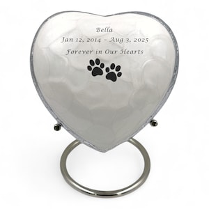 Peut inclure: Urne funéraire en forme de cœur avec une finition nacrée, soutenue par un support argenté. L'urne porte le nom "Bella", des dates et des empreintes de pattes. Le texte dit "Forever in Our Hearts".