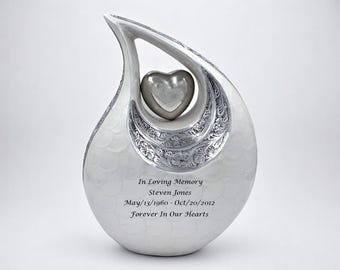 May include: A teardrop-shaped white cremation urn featuring a silver heart suspended in the center. The urn has an intricate silver floral pattern along the inner curve and is engraved with the text In Loving Memory Steven Jones May/13/1960 - Oct/20/2012 Forever In Our Hearts.