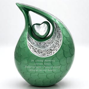 Op de afbeelding: Een traanvormige groene urn met een zilveren hartaccent en bloemmotieven. Het oppervlak heeft een schubachtig patroon en is gegraveerd met de tekst: In Loving Memory, Steven Jones, May/19/1950 - June/09/2013, Always in our hearts.