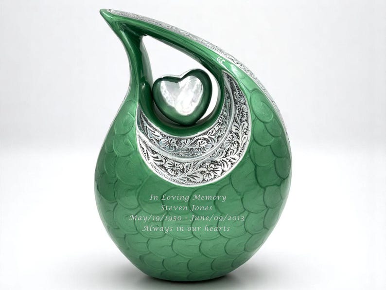 Op de afbeelding: Een traanvormige groene urn met een zilveren hartaccent en bloemmotieven. Het oppervlak heeft een schubachtig patroon en is gegraveerd met de tekst: In Loving Memory, Steven Jones, May/19/1950 - June/09/2013, Always in our hearts.