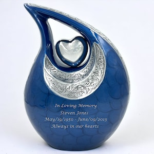 Op de afbeelding: Een traanvormige blauwe urn met een zilverkleurig hartaccent en een gegraveerde zilveren halve maanband met bloemmotieven. De tekst luidt: In Loving Memory, Steven Jones, May/19/1950 - June/09/2013, Always in our hearts.