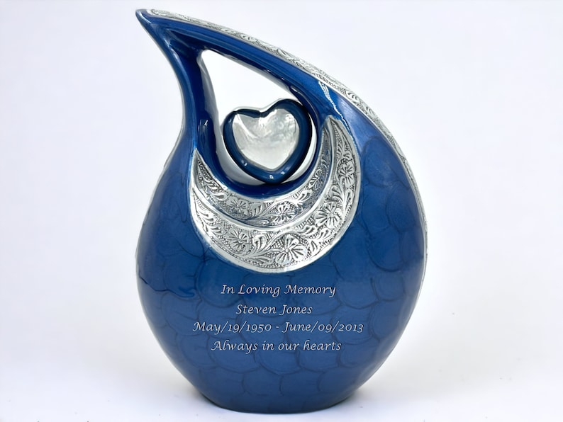 Op de afbeelding: Een traanvormige blauwe urn met een zilverkleurig hartaccent en een gegraveerde zilveren halve maanband met bloemmotieven. De tekst luidt: In Loving Memory, Steven Jones, May/19/1950 - June/09/2013, Always in our hearts.