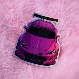May include: A holographic sticker featuring a vibrant purple sports car with a wide body kit, a prominent black rear spoiler, and a dark windscreen banner. The car is depicted from a front-facing angle, showcasing its sleek lines and headlights.
