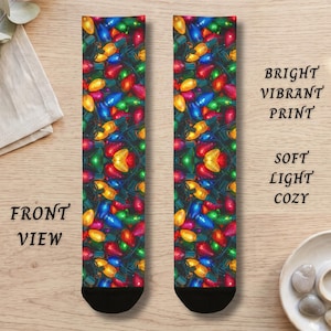 May include: A pair of crew socks featuring a vibrant, all-over print of multicolored Christmas light bulbs in red, blue, yellow, and green. The socks have black toe and heel sections and display the text BRIGHT VIBRANT PRINT SOFT LIGHT COZY.