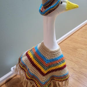 May include: A white goose statue wearing a crocheted sombrero and poncho. The sombrero is tan with blue, yellow, and red trim. The poncho is tan with stripes of blue, yellow, orange, and red, and has fringe.