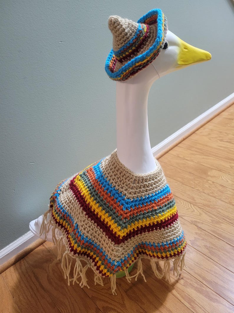 May include: A white goose statue wearing a crocheted sombrero and poncho. The sombrero is tan with blue, yellow, and red trim. The poncho is tan with stripes of blue, yellow, orange, and red, and has fringe.