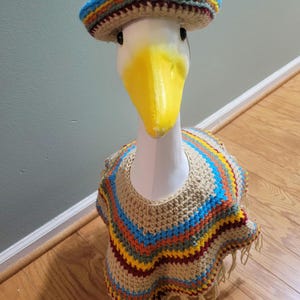 May include: A goose wearing a crocheted sombrero and poncho. The sombrero has a blue, yellow, and red striped brim. The poncho is beige with blue, orange, and red stripes and fringe.