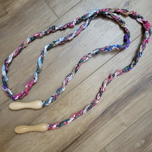 May include: Braided jump rope with a colorful fabric design. The rope features a mix of pink, blue, green, and white fabric strands. The handles are made of light-colored wood.
