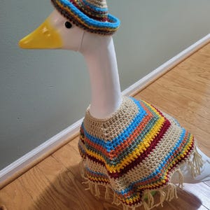May include: A white goose figurine wearing a crocheted sombrero and poncho. The hat is tan with a blue brim and colorful stripes. The poncho is tan with colorful stripes and fringe.