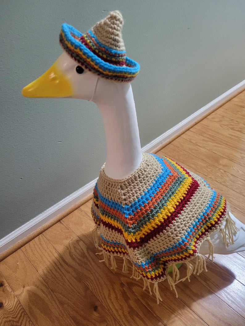 May include: A white goose figurine wearing a crocheted sombrero and poncho. The hat is tan with a blue brim and colorful stripes. The poncho is tan with colorful stripes and fringe.