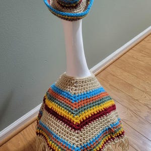 May include: A crocheted sombrero and poncho set. The hat has a blue brim and the poncho features a striped pattern in blue, yellow, orange, and red. The set is beige with fringe.