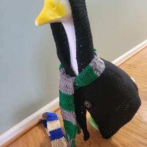 May include: A decorative goose head with a yellow beak, wearing a black crocheted hooded coat and a green, gray, and white striped scarf. A blue and tan striped scarf lies on the floor. The goose is on a wooden surface.