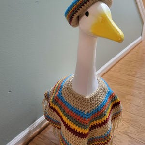 May include: A decorative goose head wearing a crocheted sombrero and poncho. The hat has a blue, yellow, and red striped brim. The poncho is beige with colorful stripes and fringe.