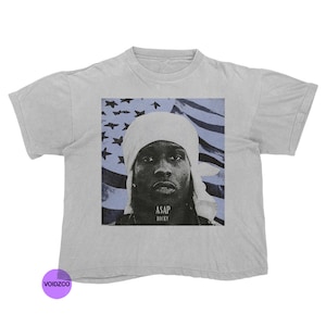 Puede incluir: Camiseta gris claro con un gráfico de una persona con un tocado blanco, sobre un fondo de un diseño de bandera americana azul. El texto "ASAP ROCKY" está impreso debajo de la imagen.