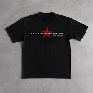 Pode incluir: Camiseta preta com o nome da banda "Rage Against The Machine" em texto branco e uma estrela vermelha. O texto "killing in the name" aparece abaixo do nome da banda em branco.