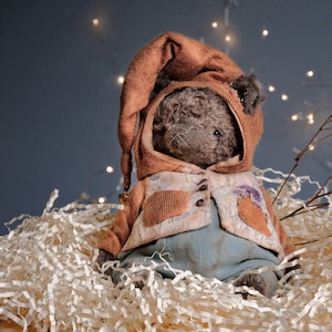 May include: A handmade teddy bear with a brown hooded cap and a matching vest. The vest has a floral pattern and is buttoned with three small, dark buttons. The bear is sitting on a bed of white paper strips.