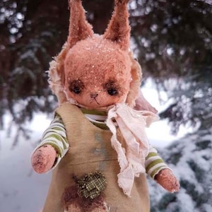 May include: A handmade plush toy squirrel with long ears, wearing a green and white striped shirt and a brown and green patched overall. The squirrel is holding a white fabric scarf in its hands.