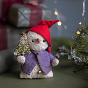 May include: A white plush toy snowman wearing a purple jacket and a red hat, holding a small green Christmas tree. The snowman is standing on a green surface.