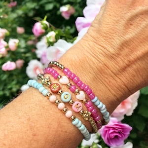 May include: A set of five bracelets with pink, blue, and gold beads. The bracelets spell out "MOM" with gold letters. The bracelets are stacked on a person's wrist.