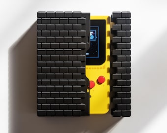 May include: A yellow handheld video game console with a black and white screen and two red buttons. The console is partially enclosed in a black case with a brick pattern.