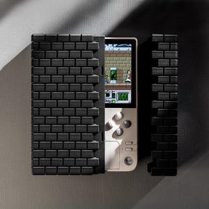 May include: A white handheld gaming console with a black brick pattern on the front and back. The console has a screen showing a video game with a green and brown background. The console has a white button with the letter B on it.