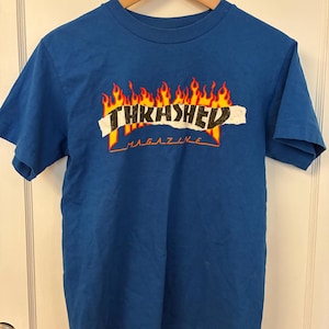 May include: A bright blue short-sleeve t-shirt featuring a graphic print of orange and yellow flames behind a white torn paper strip with the black text THKASHED and the word MAGAZINE printed in orange underneath the flames.